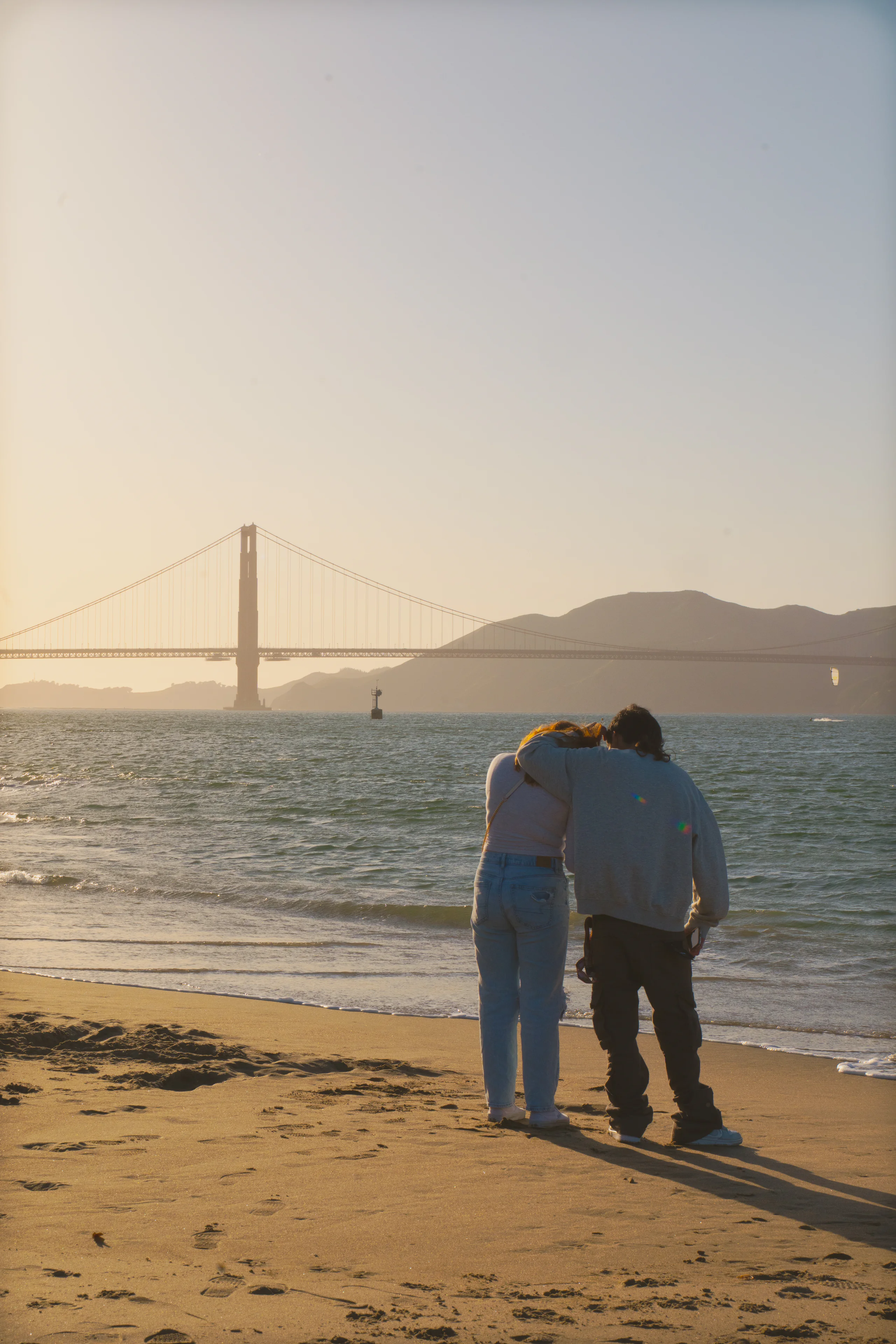 A couple embraces on a beach with a suspension bridge across the water behind them.