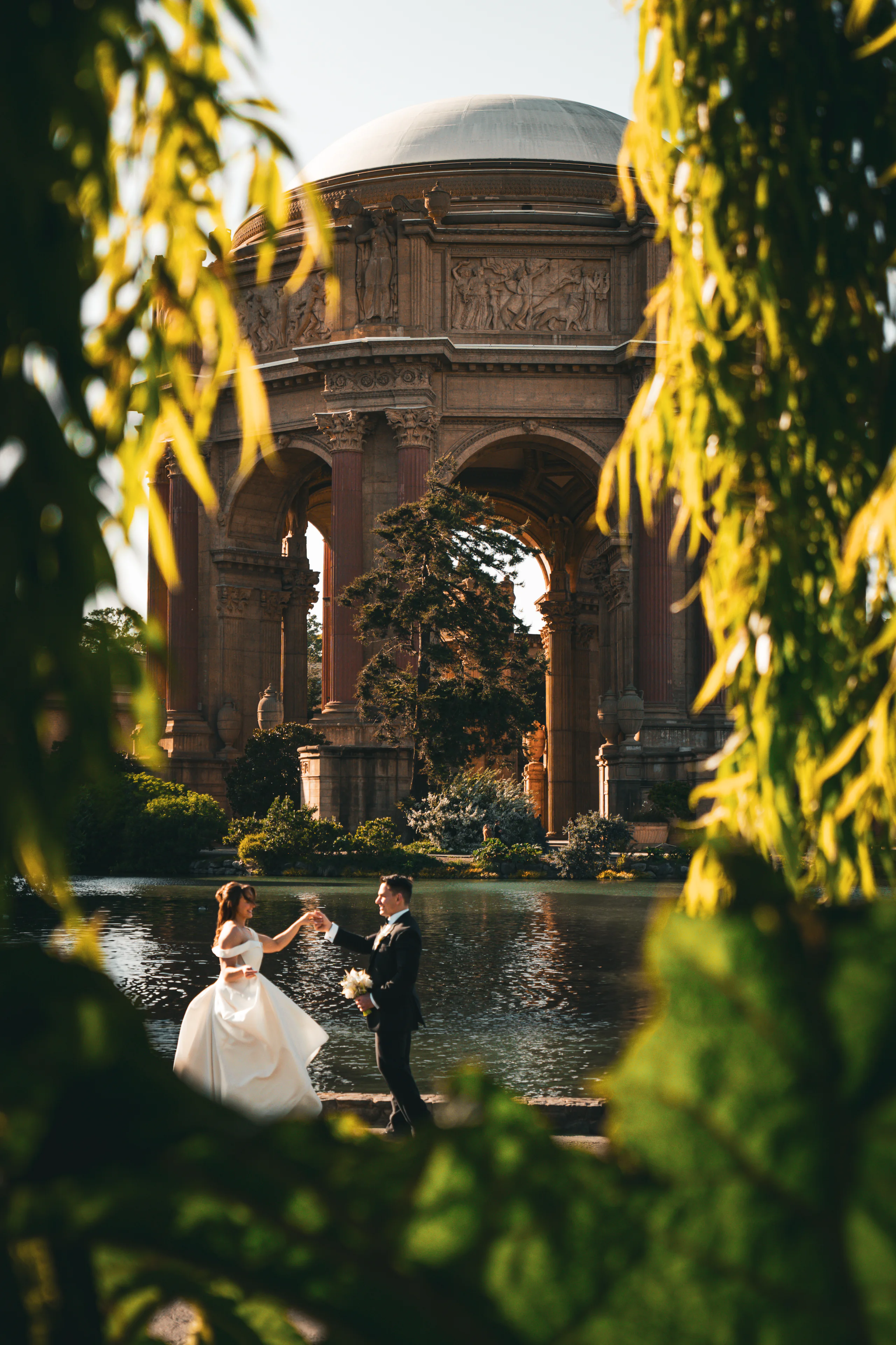 A wedding couple dances beside a reflecting pool, framed by hanging leaves and domed architecture.