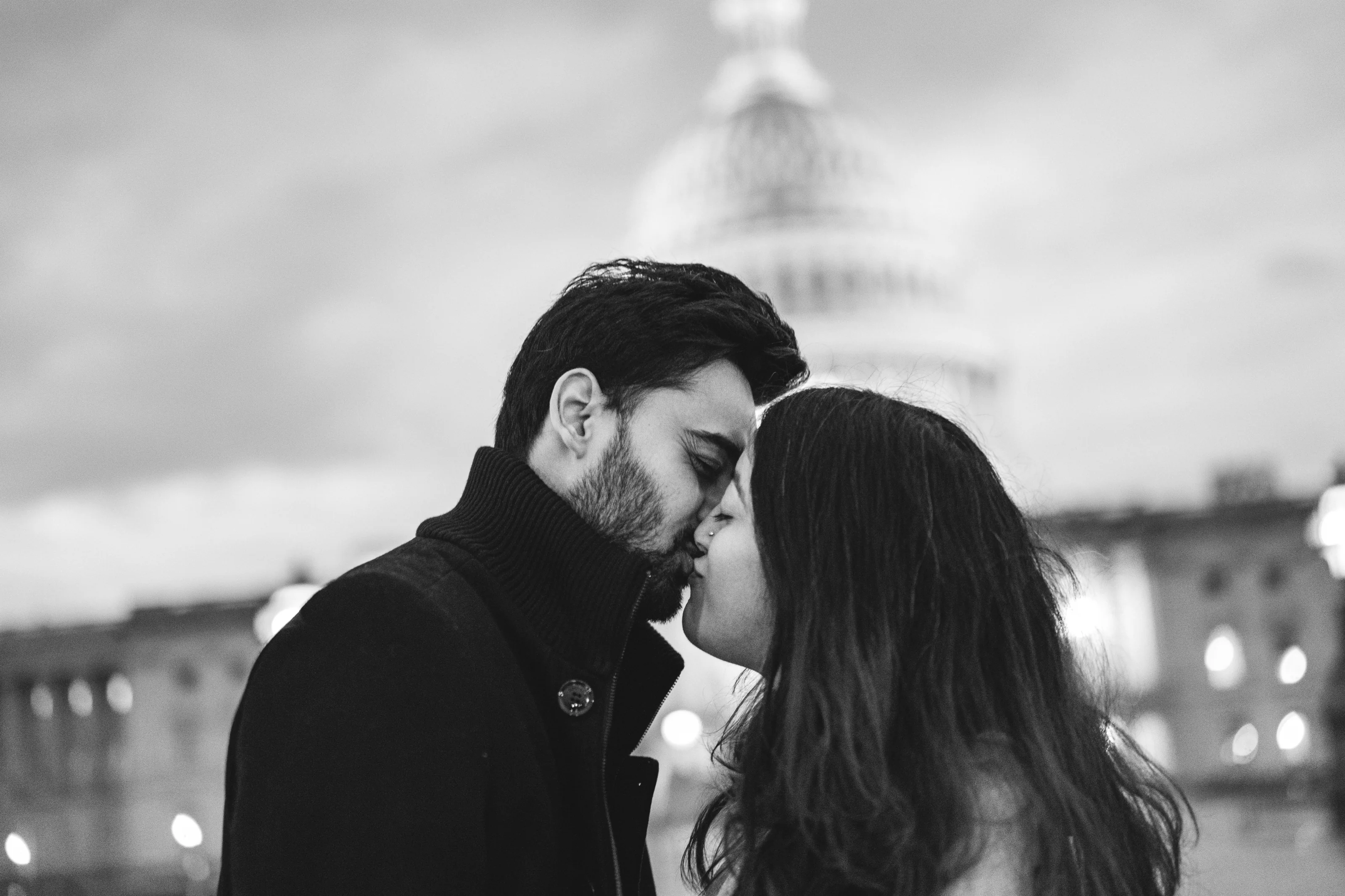 A couple shares a kiss in black and white with a domed building softly blurred behind them.