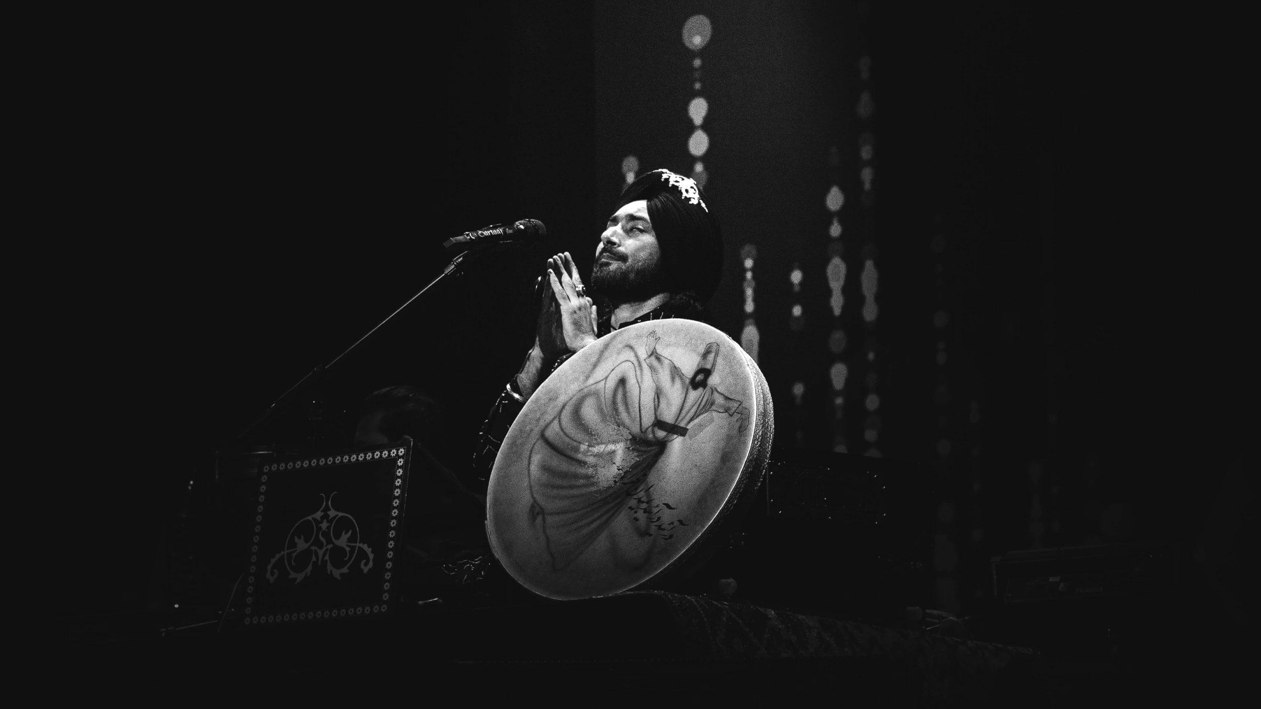 A musician in a turban holds a hand drum beside a microphone on a dark stage.