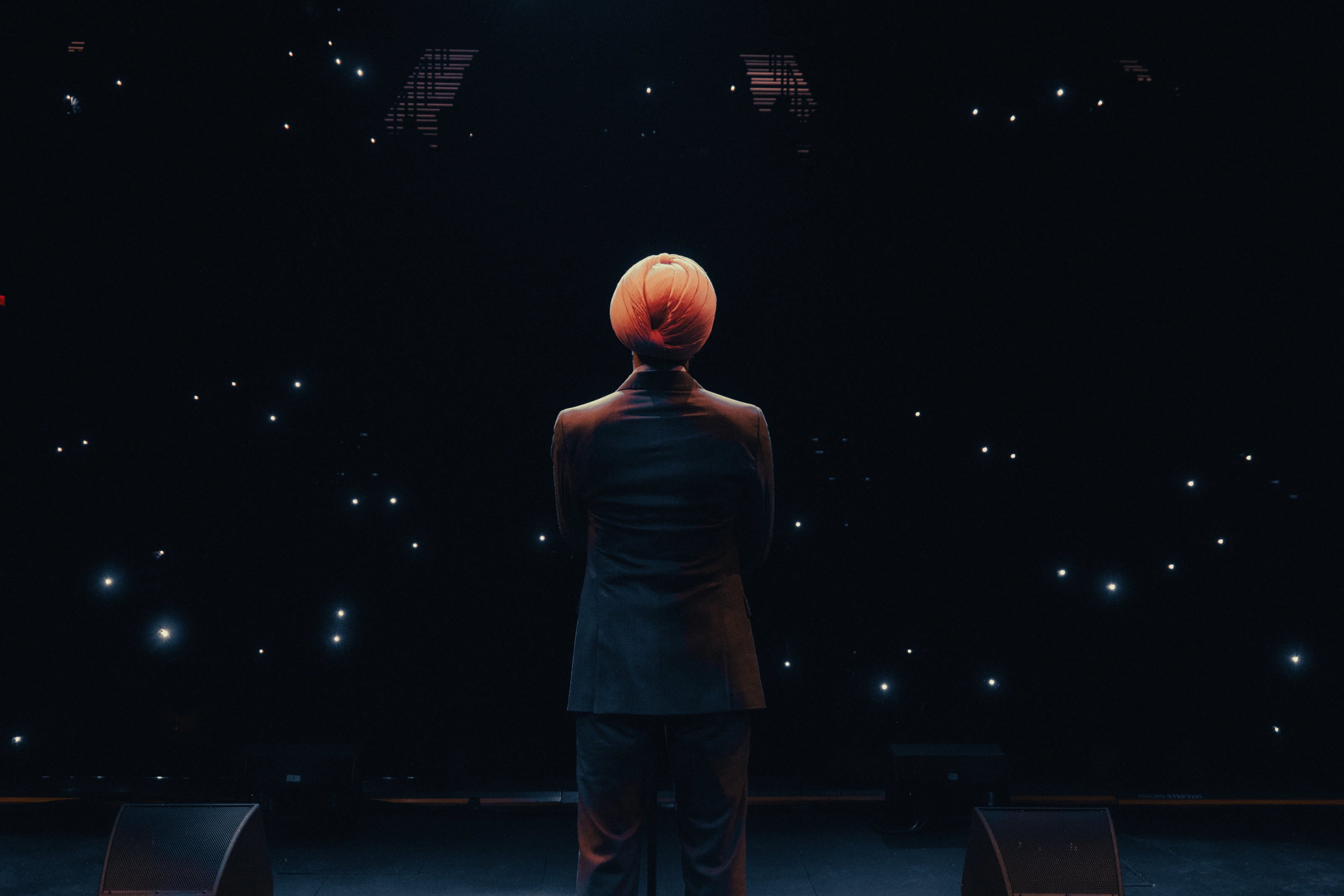A performer faces an audience from a dark stage lit by phone lights.