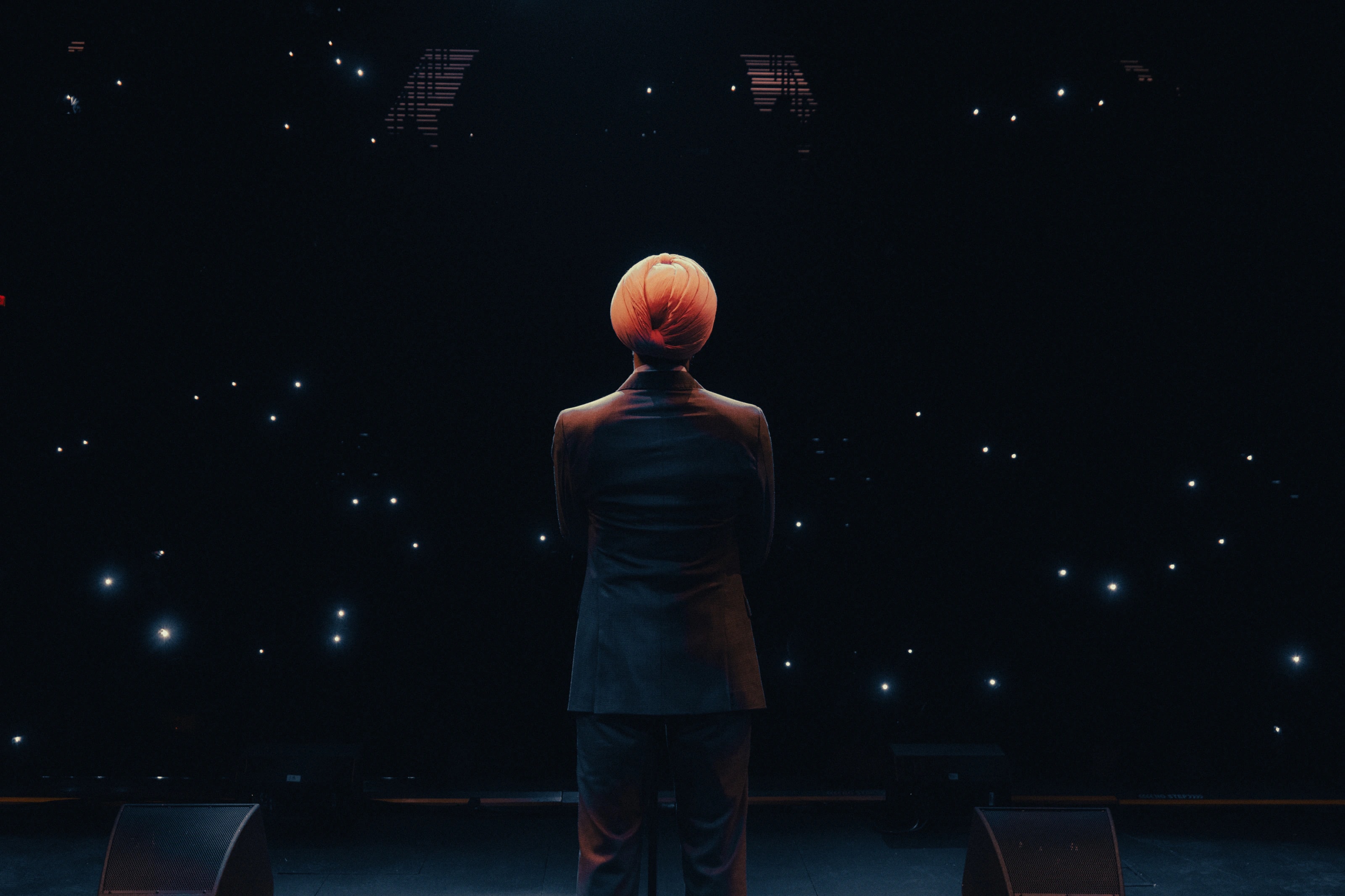 A performer in a suit and turban faces an audience from a dark stage lit by phone lights.