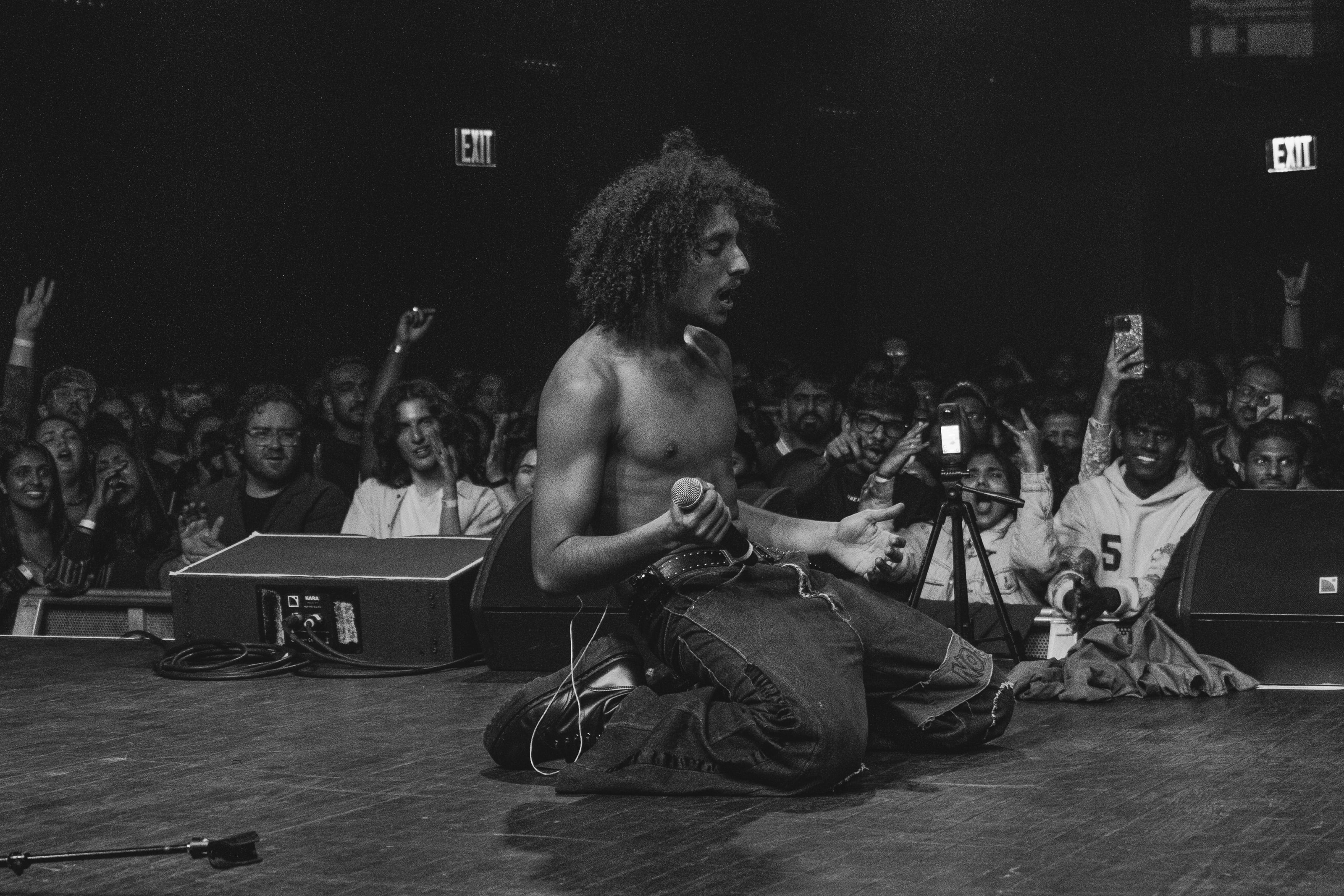 A performer holding a microphone sits at the edge of a stage in front of an engaged audience.